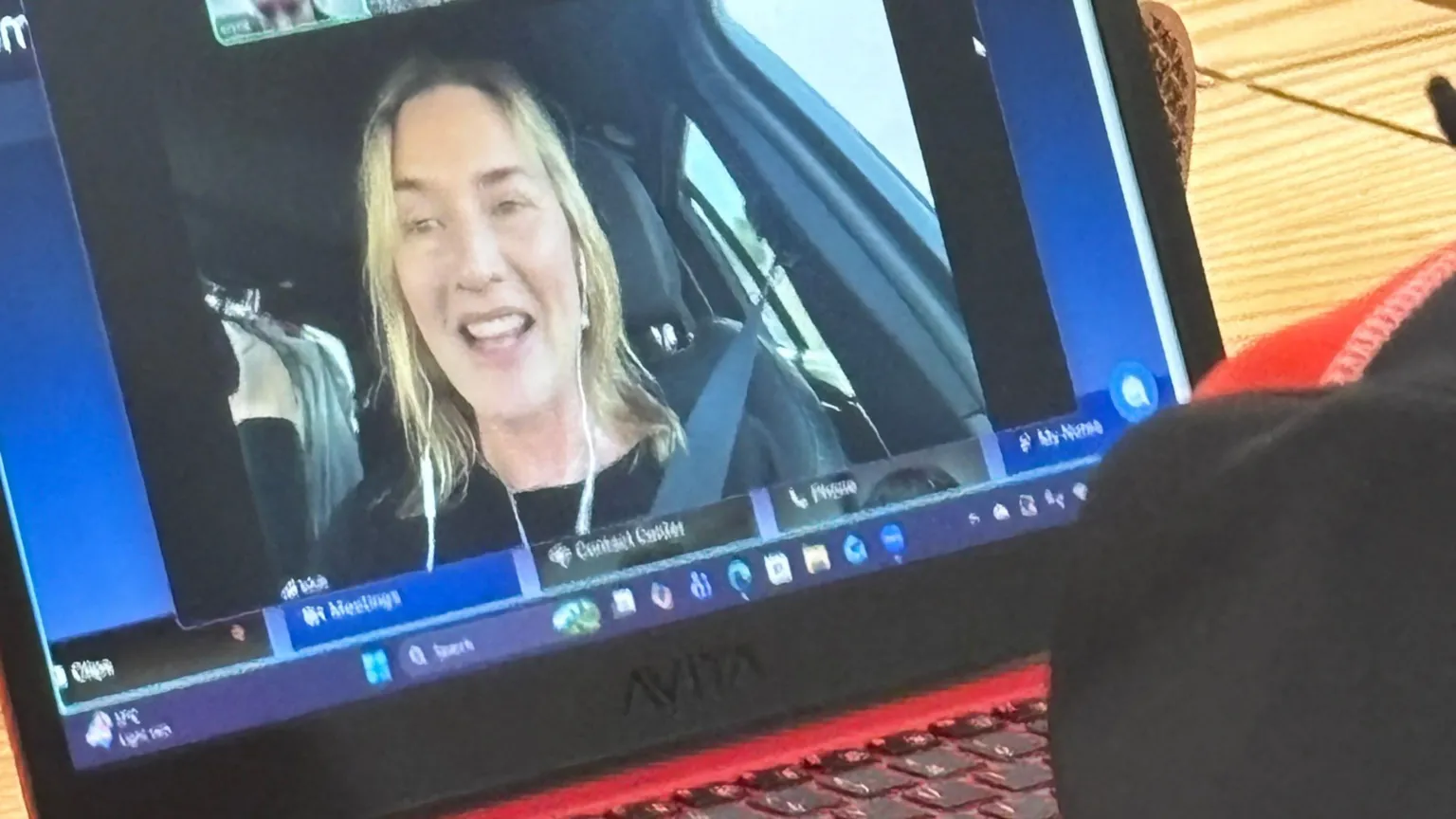 A close up image of a laptop screen. A video call is underway and the screen is filled with the picture of a woman talking. She is sitting in a car with her seatbelt on. She has white wired earphones in and has a big bright smile. Her hair is long and blonde and she is wearing a black top. 