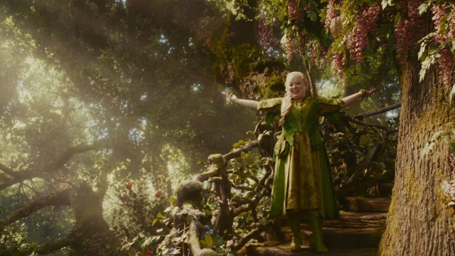 NEAL STREET PRODUCTIONS A still image from The Magical Faraway Tree film. It's a forest scene with a tree closest to us at one side. Standing on stairs to the tree is a woodland fairy. She is dressed in layers of green clothes with boots and a bag. Her arms are outstretched and she's smiling in a welcoming way. Behind her are trees and flowers with a beam of sunlight making everything glow. 