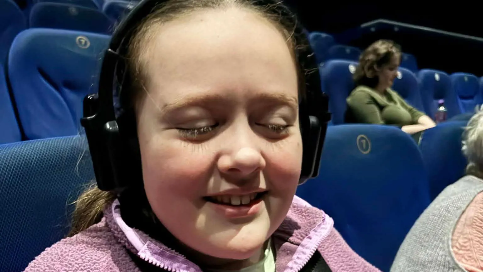 Evanna Kirkpatrick A girl is in a cinema, with rows of chairs all around her. Her eyes are closed and she is smiling. She's wearing a pink fluffy fleece with a zip and her long dark hair is tied back. She is wearing a pair of black headphones which are for audio describing the film. 