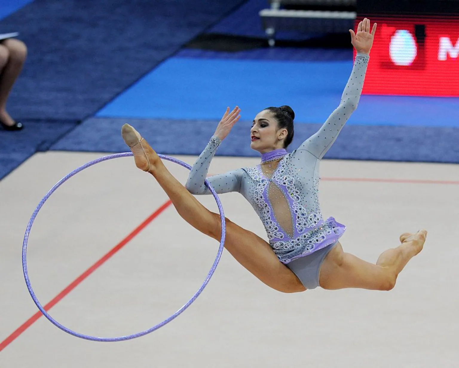 AFP via Zeynab Javadli at a gymnastics competition in 2011. She is wearing a blue and purple leotard embellished with jewels. The photo captures her jumping in the air doing a split kick with her front leg and arm going through a large hoop.