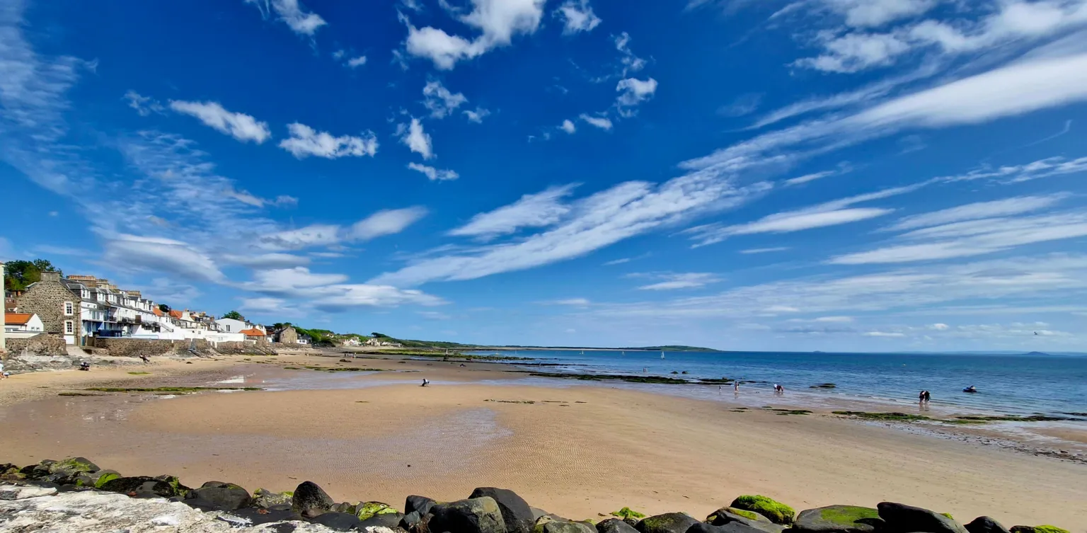 Erin Harper Houses look across the beach at Lower Largo. There are few clouds in the sky and people walking along the beach. The sea is bright blue and rocks are at bottom of the image.