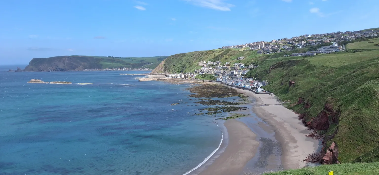 Jim Mclean Two towns on the side of the cliffs. The sea and the beach are at the bottom of the rocks which are covered in grass. The sky is clear with no clouds. 