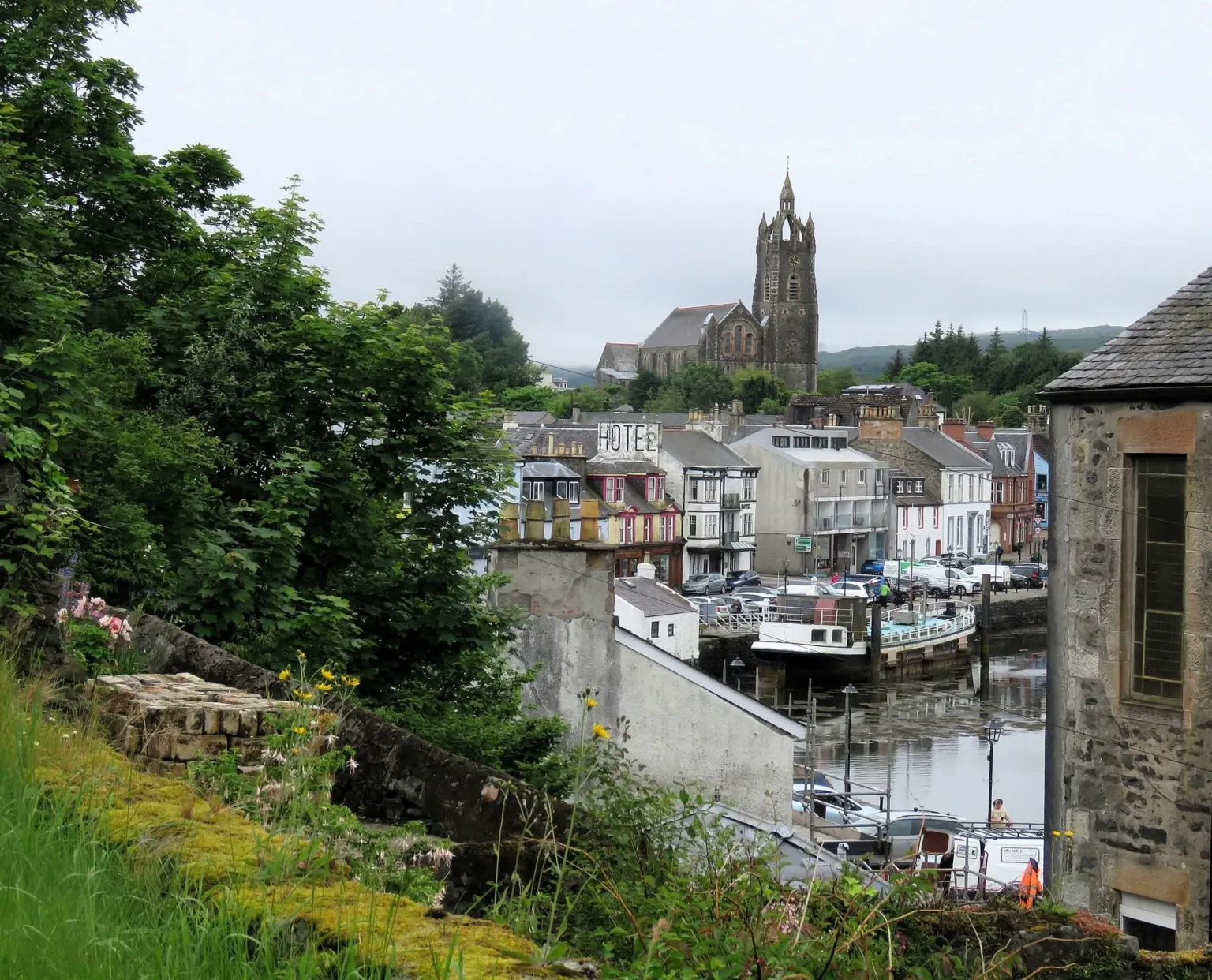 Rachel Hall A street of houses overlooking a harbour. A church stands on top of a hill in the background and there are trees and bushes in the foreground.