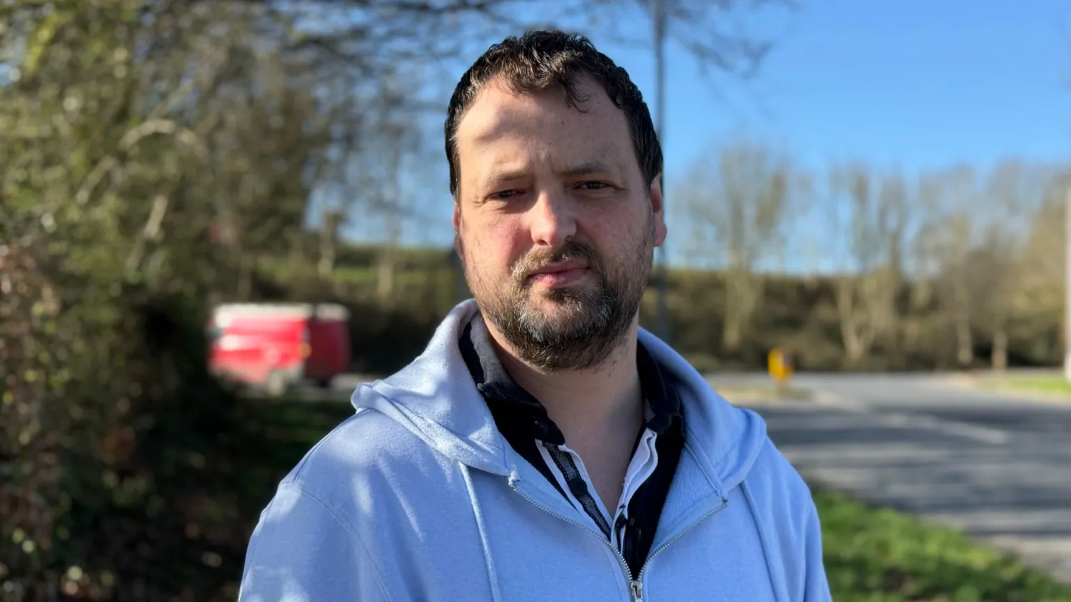 Malachy Quinn has short dark hair and a dark beard. He is standing outside on a grassy area with bushes behind him. He's wearing a light blue zip up hoody and navy polo shirt. 