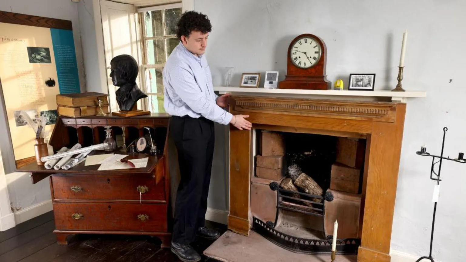 Colin Hattersley A man wearing a blue shirt and black trousers points to the wooden mantelpiece which has picture frames, a clock and candlestick on top of it. To the side is a wooden writing desk adorned with letters and books. 