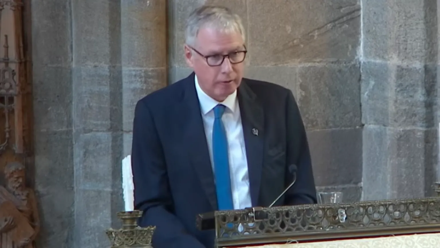 Dunblane Cathedral A man with short grey hair and glasses is wearing a black jacket, white shirt and blue tie as he stands at a church lectern
