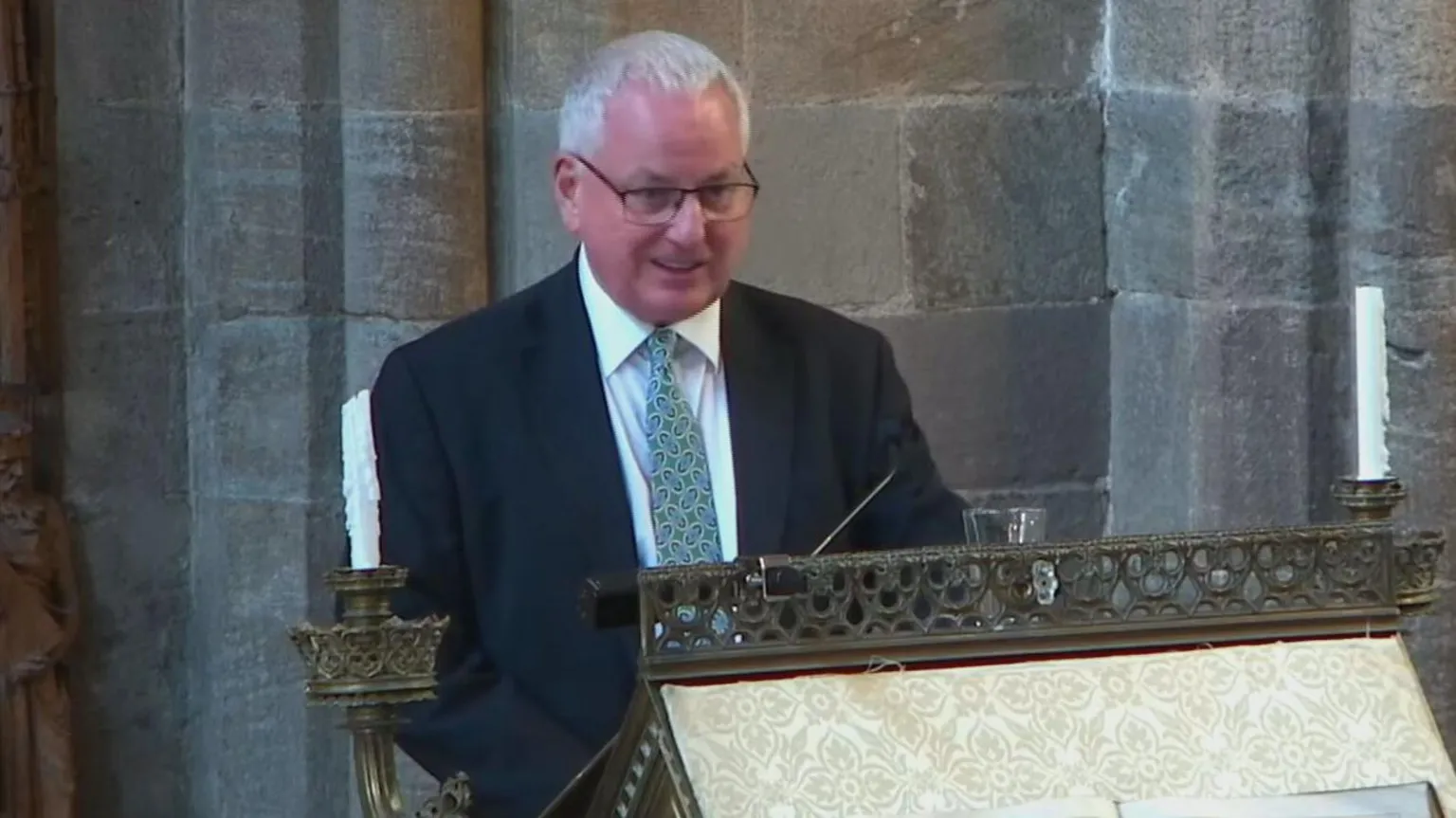 Dunblane Cathedral A man with short grey hair and glasses is wearing a black jacket, white shirt and green tie as he stands at a church lectern