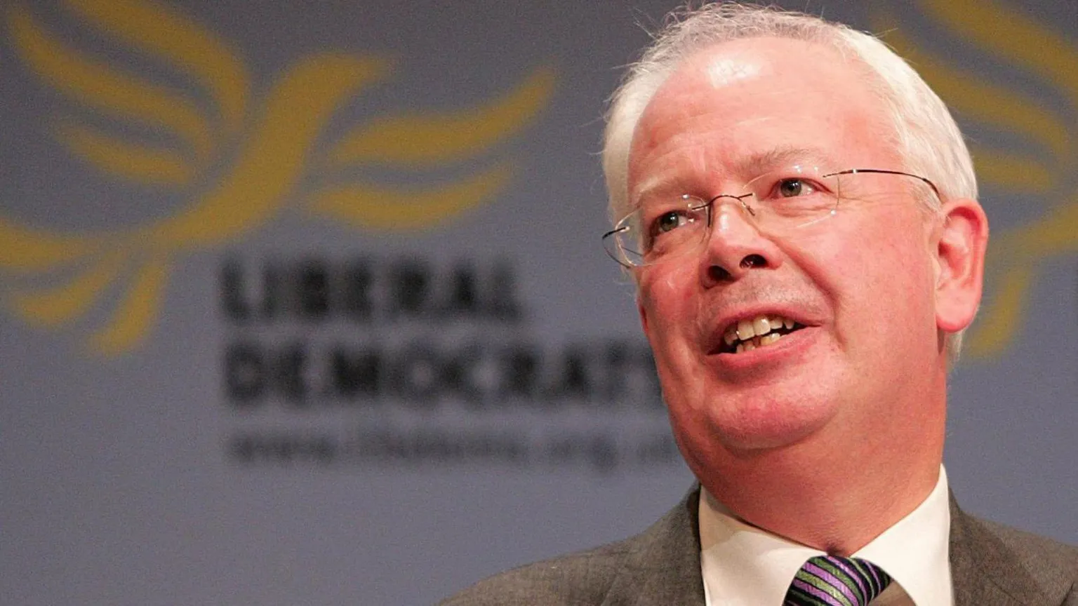  Jim Wallace in striped tie and grey suit in front of a party sign.