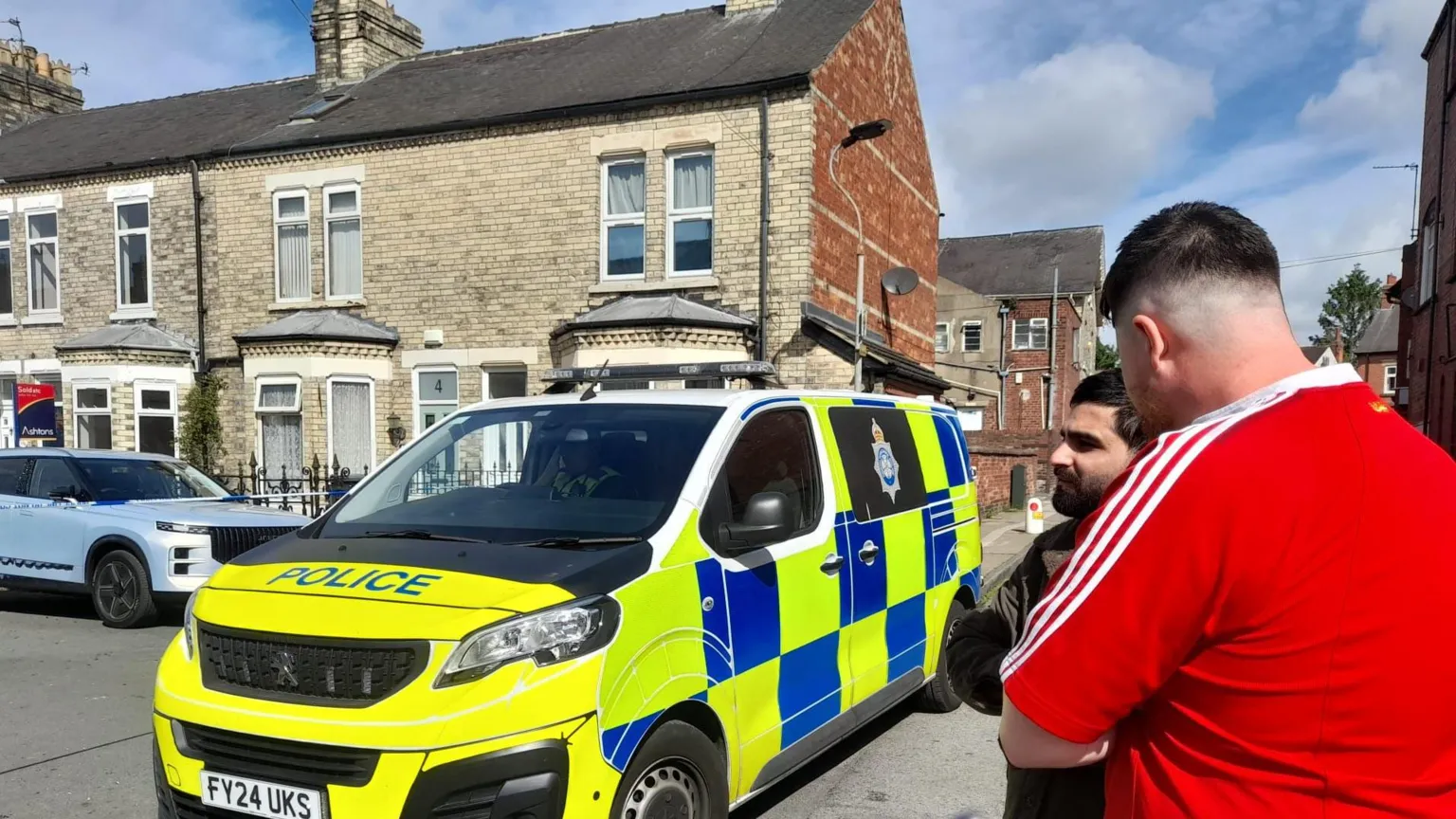 BBC/Andrew Barton The image shows a residential street scene in daylight. A police van is parked on the road outside a row of terraced houses. On the right side of the image, two people are standing close together near the police vehicle. One man is wearing a bright red short-sleeved top with white stripes on the shoulders and light grey trousers. The other man, partially turned away, is wearing darker clothing.