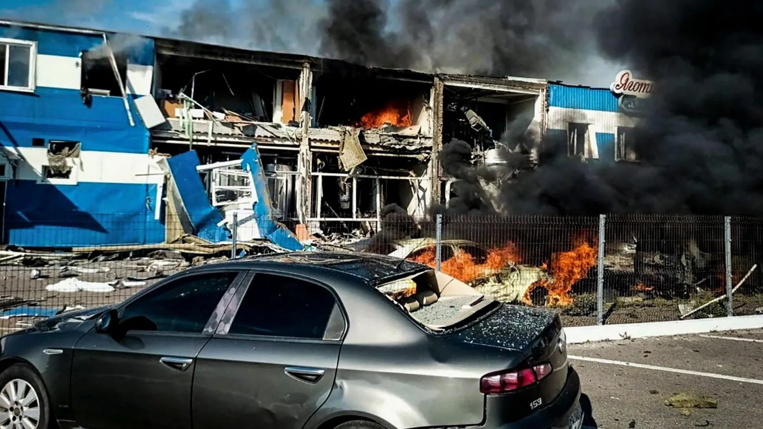 EPA/Shutterstock Black smoke is seen rising from a building, where some of the rooms are exposed and flames can be seen inside. A car is on fire in front and another car is seen in the foreground. 