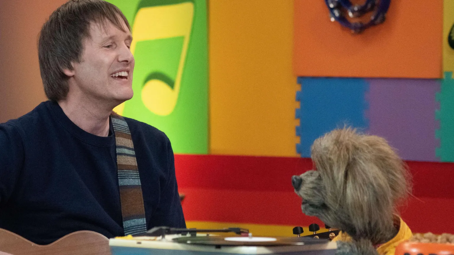 Tim and the CBeebies dog Dodge sitting together singing. They're sitting in a multi-coloured room and Tim is holding a guitar. He's wearing a navy top.