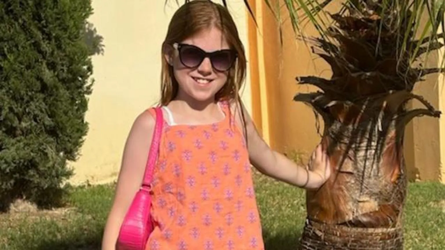 Supplied Edie Smart is smiling on a sunny day leaning against what looks like a palm tree. She is wearing an orange, sleeveless top and has a pink bag over her shoulder. She is wearing big sunglasses and has fair, long hair. 