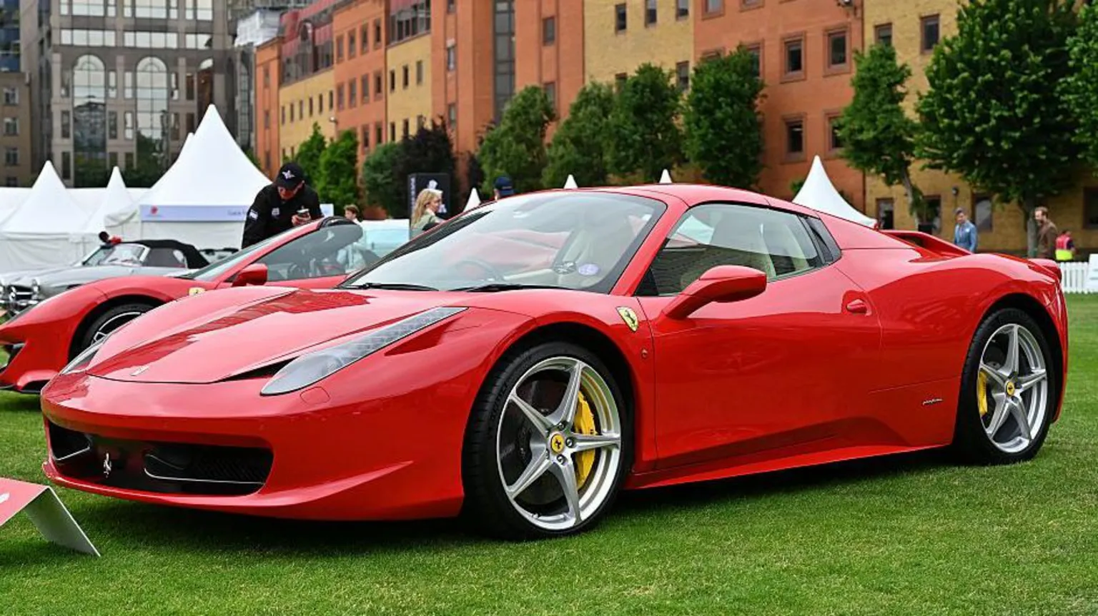  A red ferrari sports car parks on a lawn with more another Ferrari and a Mercedes in the background as well as brick buildings in London