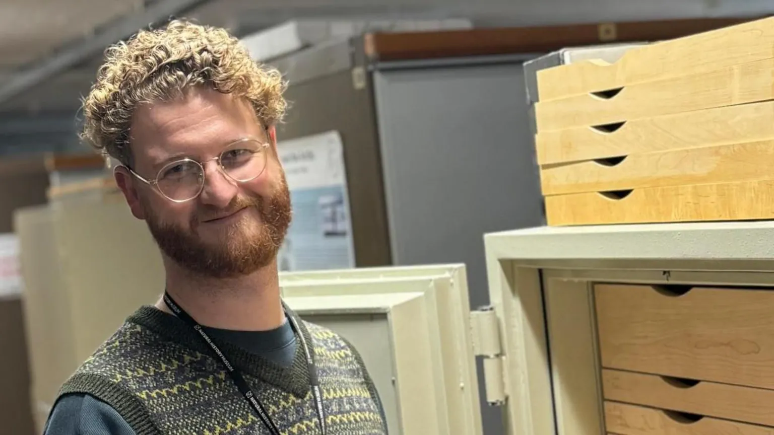 Richard Howard Richard looking at camera smiling. He stands next to a filing cabinet and wears a grey t-shirt, a patterned waistcoat and clear round glasses. He has curly blonde hair. 