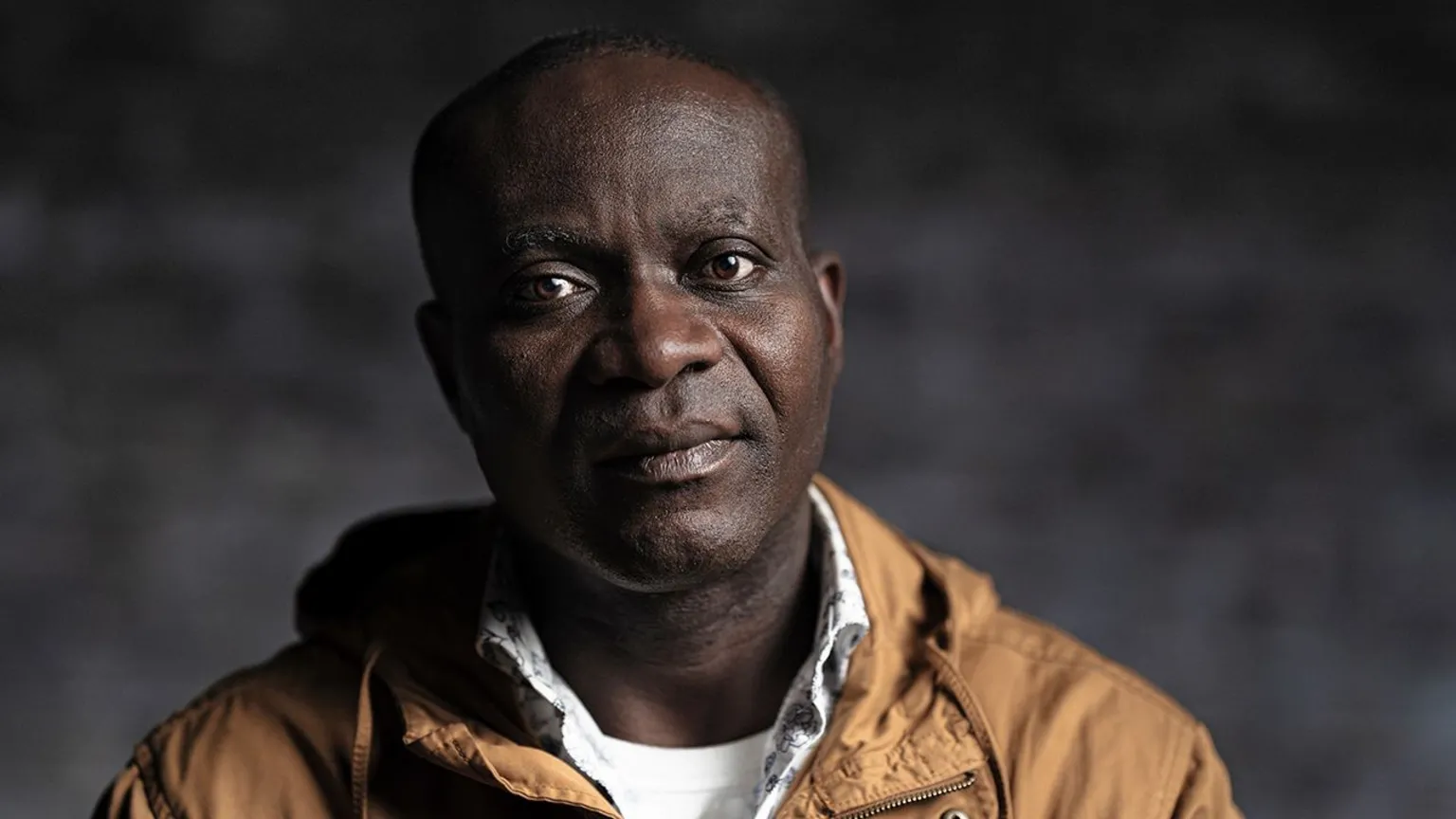 Gavin Hopkins Augustus Mensah is bald and looks at the camera with a neutral expression. He wears a brown jacket and white t-shirt