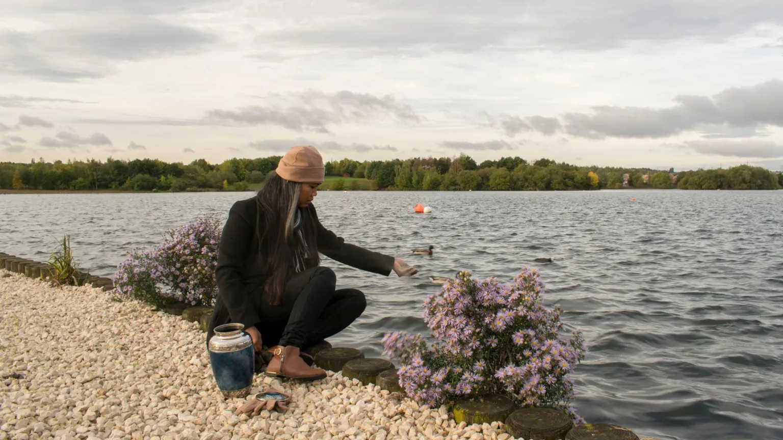  Woman at side of water on pebble stone path and by water and with urn by her side scattering ashes