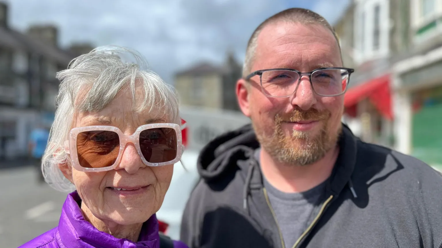 On the left is an older woman with short grey hair and pink framed sunglasses on. She's smiling and wearing a purple coat. Next to her is a man with short grey hair and a grey and ginger beard. He's wearing glasses, a grey t-shirt and grey hoodie. 