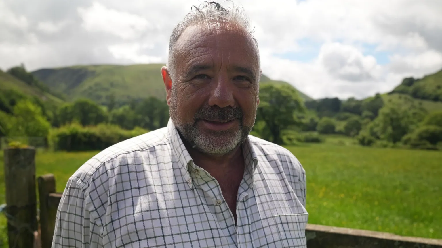 A man smiling at the camera. He has short grey hair, a grey beard and is wearing a white and black check shirt