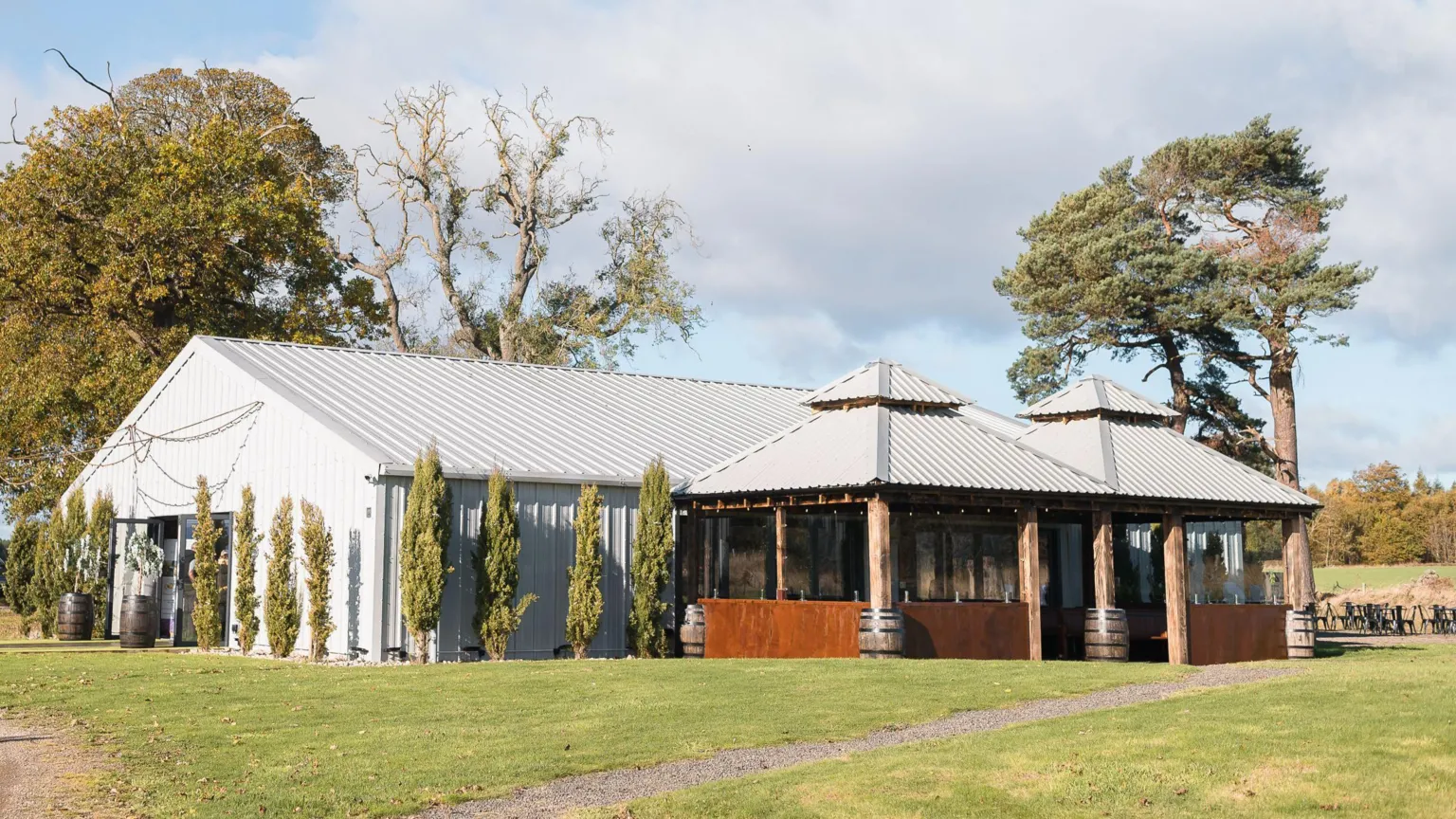 A large barn made of steel and wood sits in a large green expanse, surrounded by trees 