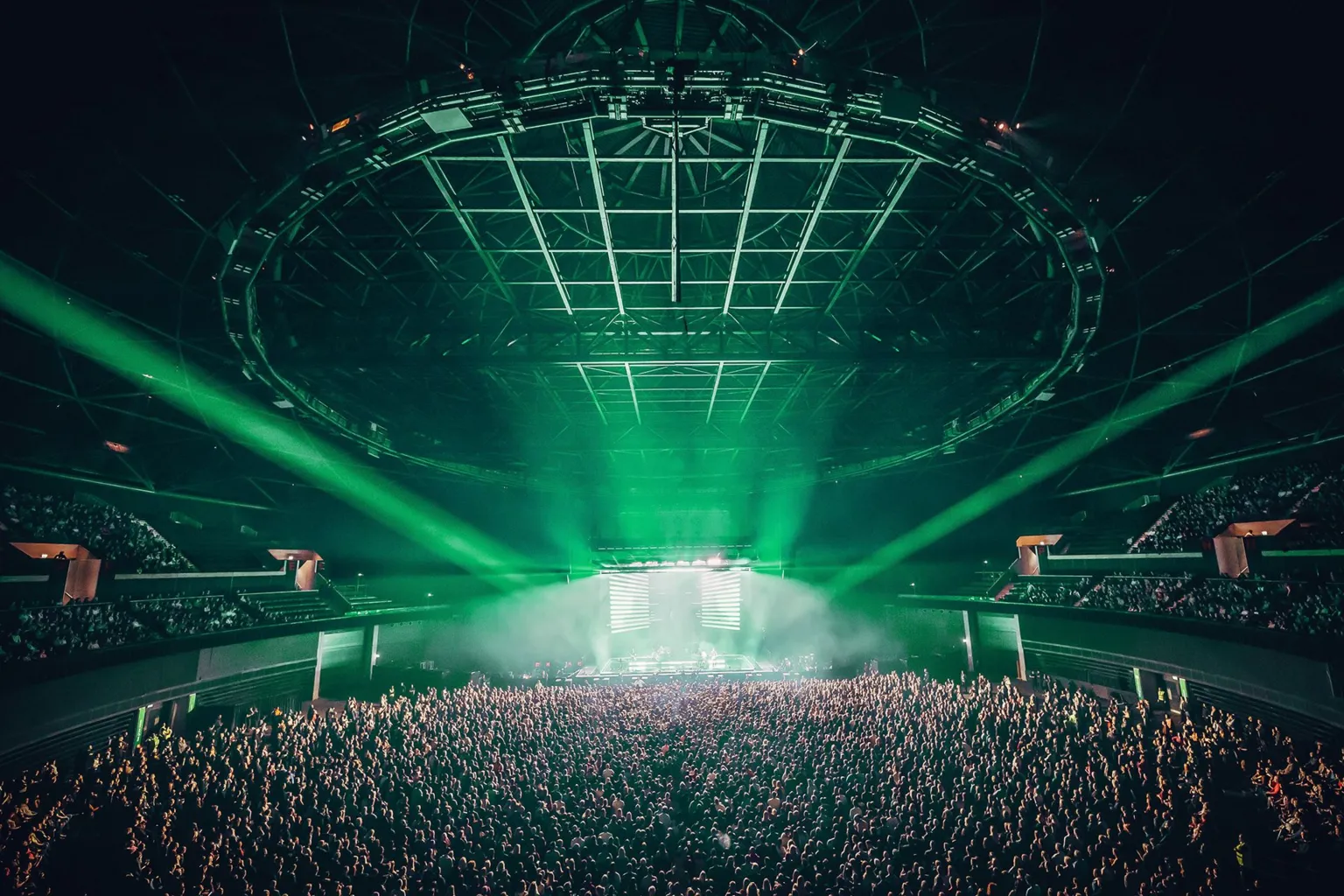 Hydro Inside shot of the area showing its vast space. Thousands of fan stand in the pit and in the seating to the sides. Green lights shine on the stage. 