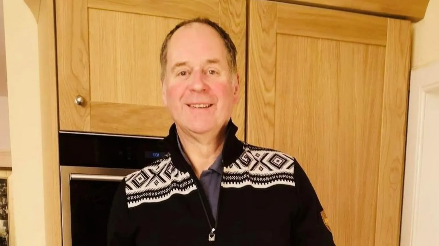 Crowley family David Crowley, a man smiling at the camera, he is standing inside a house, and is wearing dark jumper and trousers.