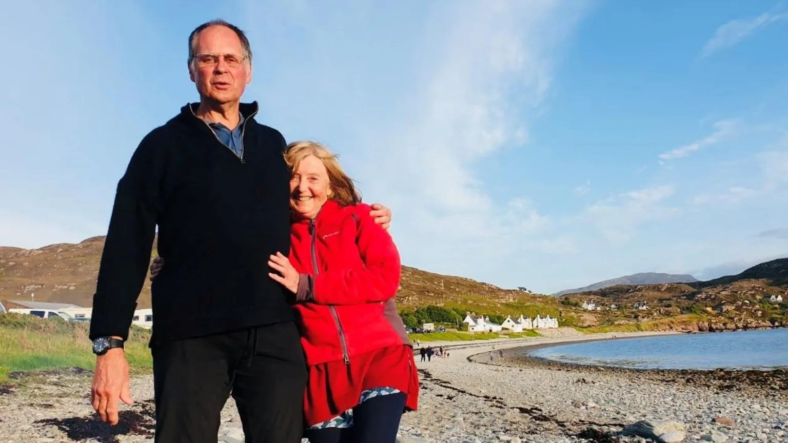 Crowley family David and Mary Crowley, he is wearing a dark jumper and trousers, she has on a red jacket, they are smiling at the camera, while standing on a shore next to water.