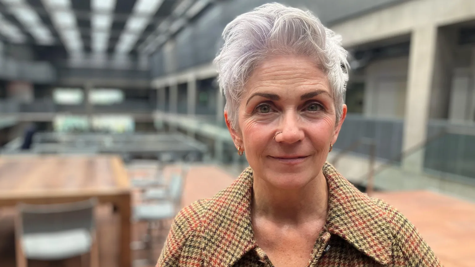 Denise Mina, a woman with short grey hair, is wearing a tweed jacket with a black shirt underneath. She is smiling, standing in a grey modern building with table and chairs in the background.