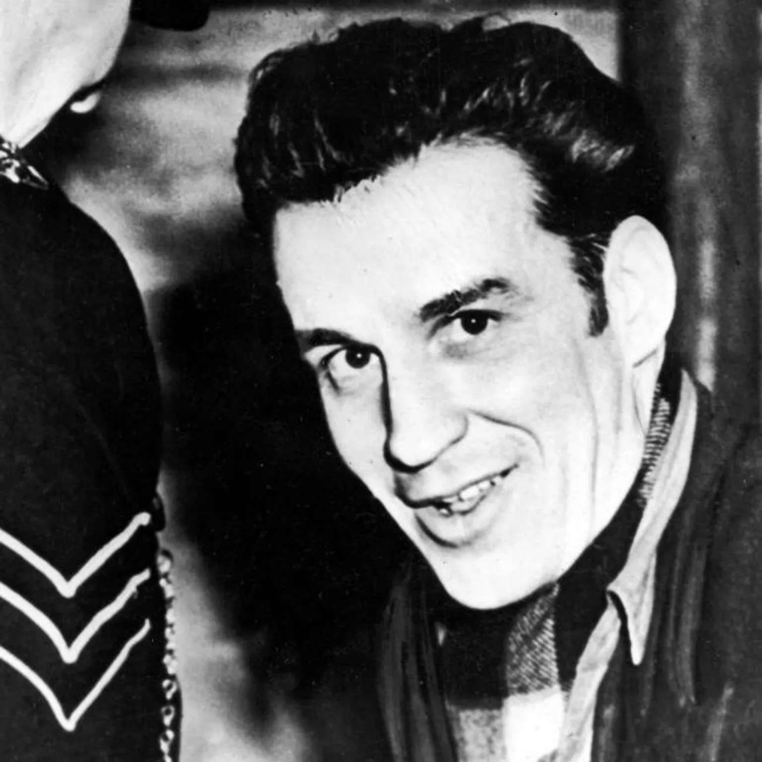  Manuel, with thick dark hair and long sideburns, smiles at the camera. He is dressed in a dark jacket, open necked casual shirt and tartan scarf. A police sergeant is facing him, away from the camera.