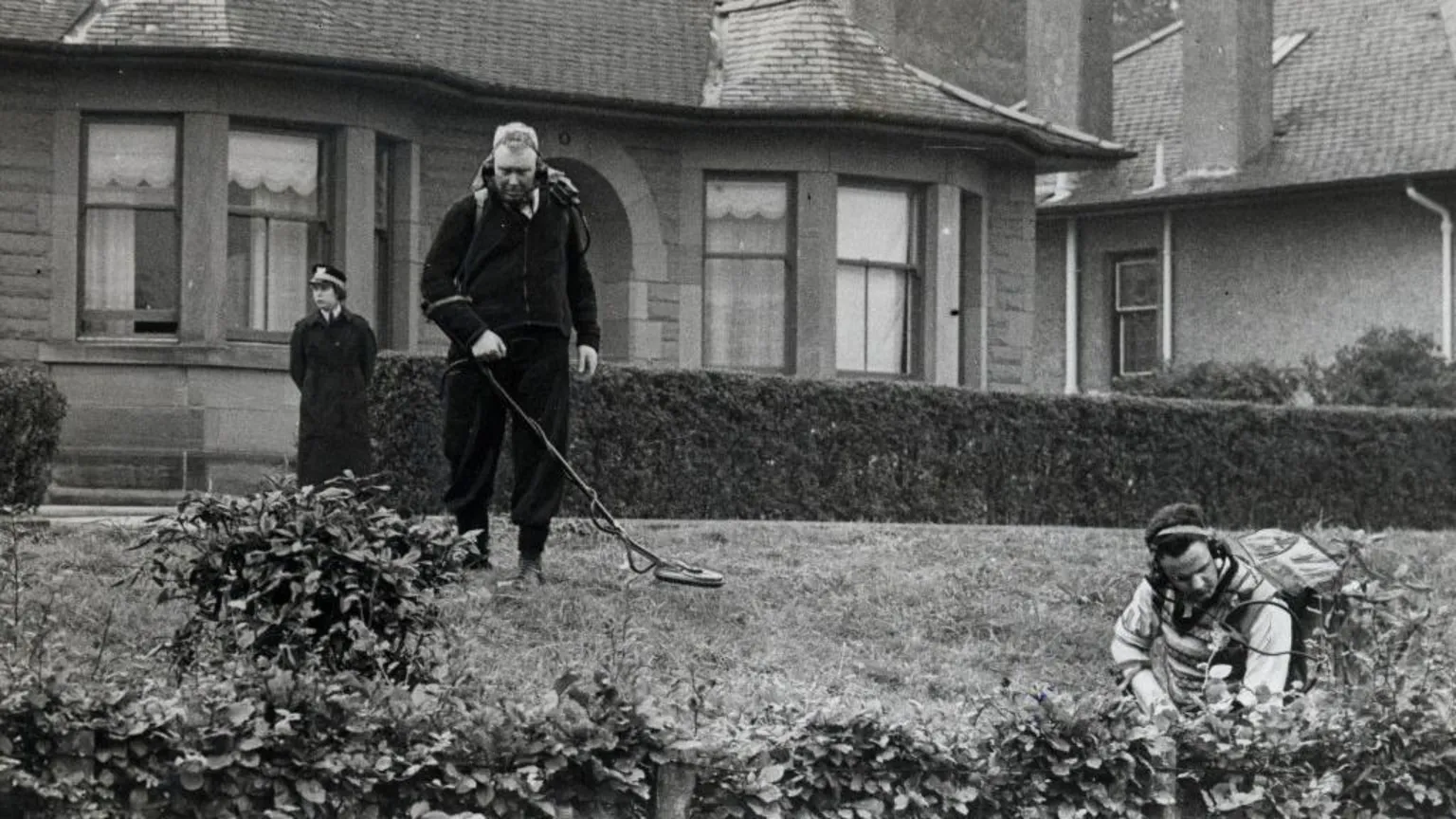  Three police officers, two with metal detectors, are searching some ground and bushes outside a stone bungalow.