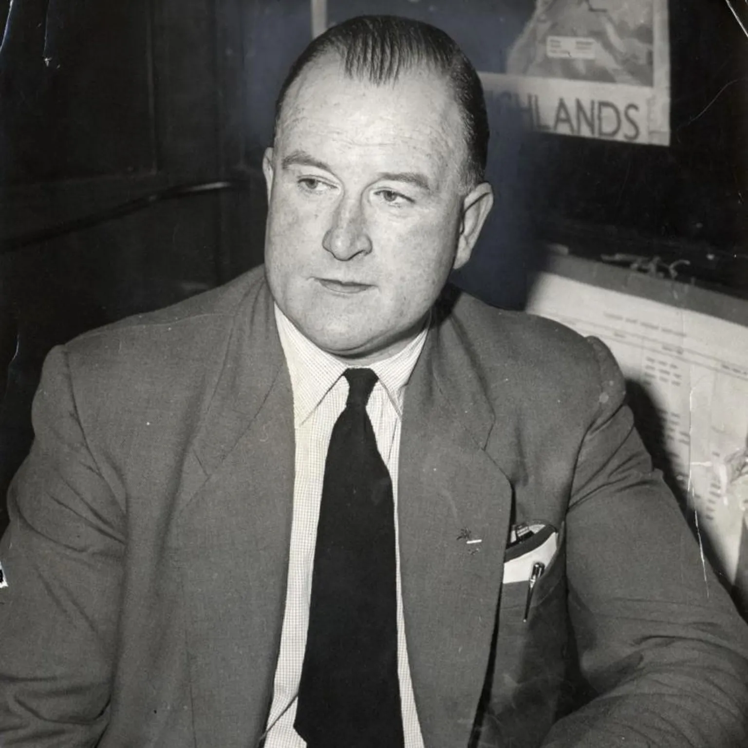  William Watt is a balding man in a grey suit with white shirt and dark tie. He is sitting, looking off-camera.
