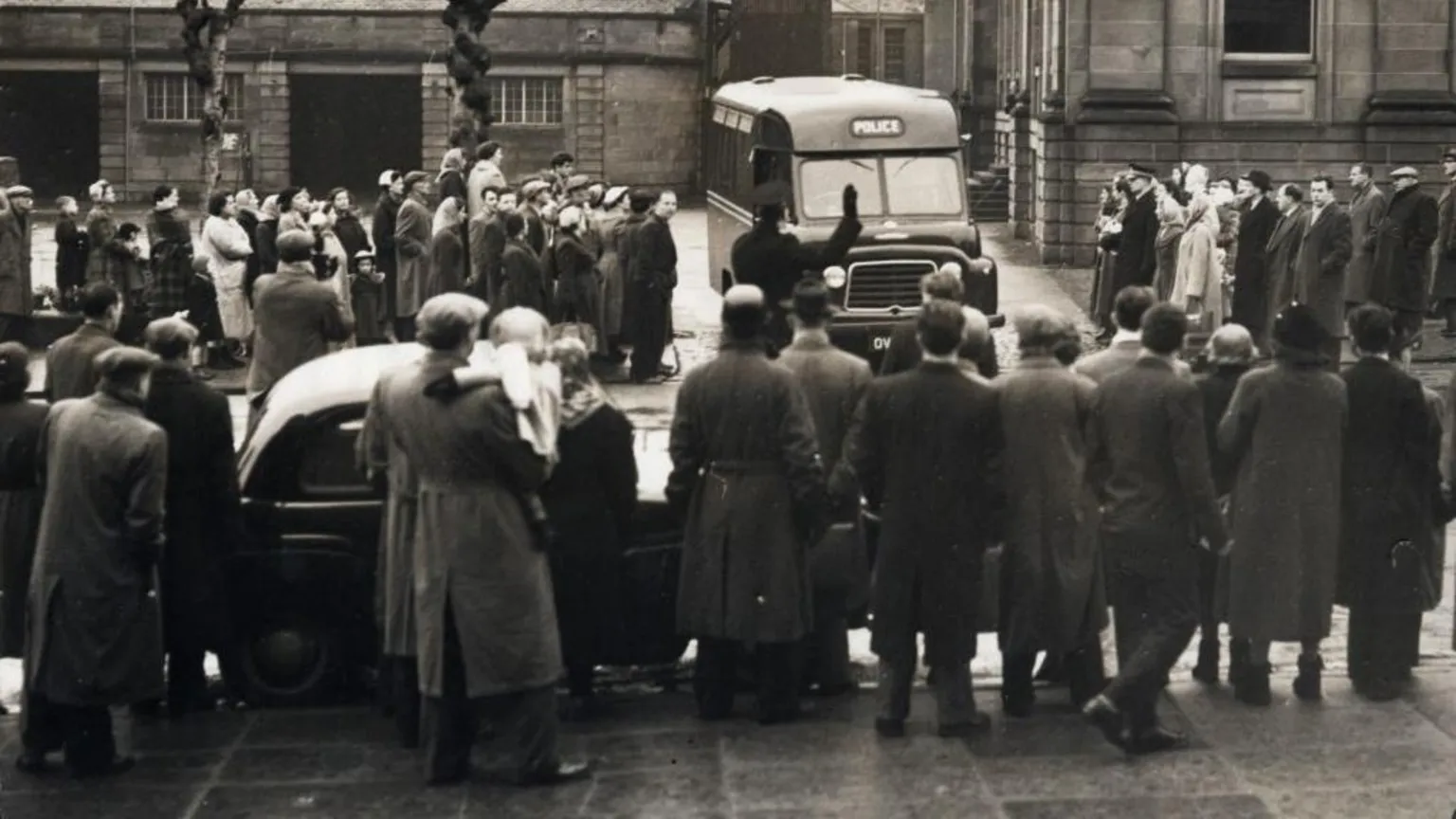  A large crowd of onlookers stand outside the court building. A prison van is moving towards the crowd and a police officer can be seen holding his hand up in front of it. 