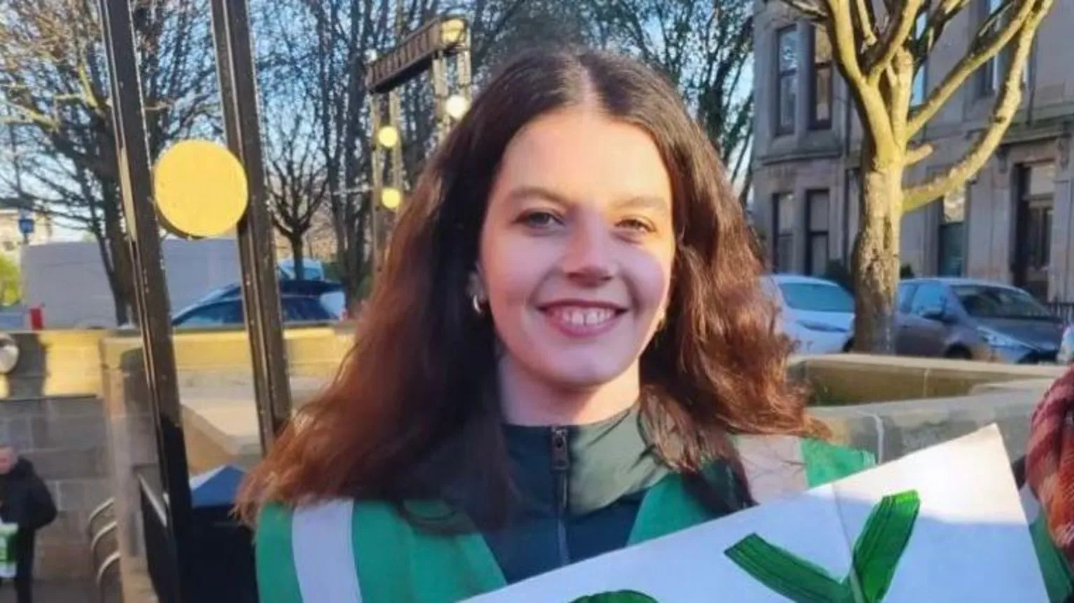 Stella Rooney A woman with long dark hair, holding a placard while out on a street. 