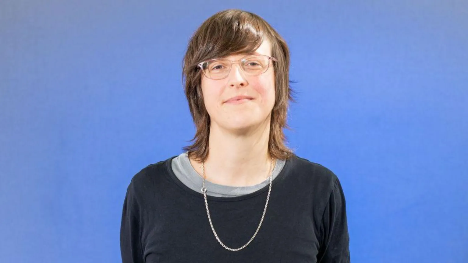  A woman with shoulder length brown hair stands in front of a blue background. She is wearing a blue sweater, a long necklace and glasses. 