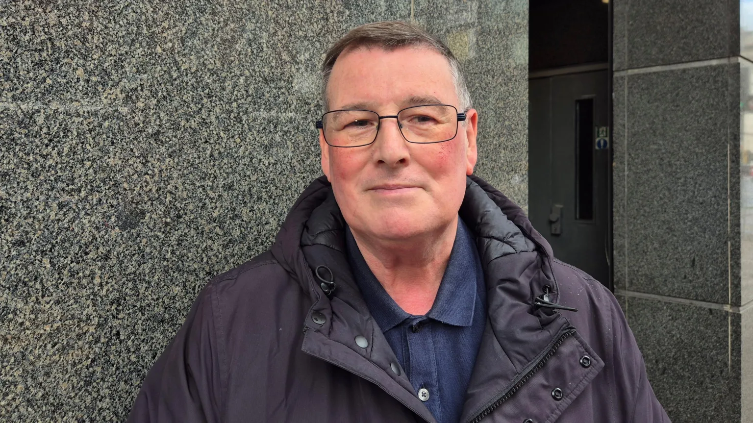 A man wearing a dark jacket, with glasses and short, dark and greying hair. He is standing outside. 