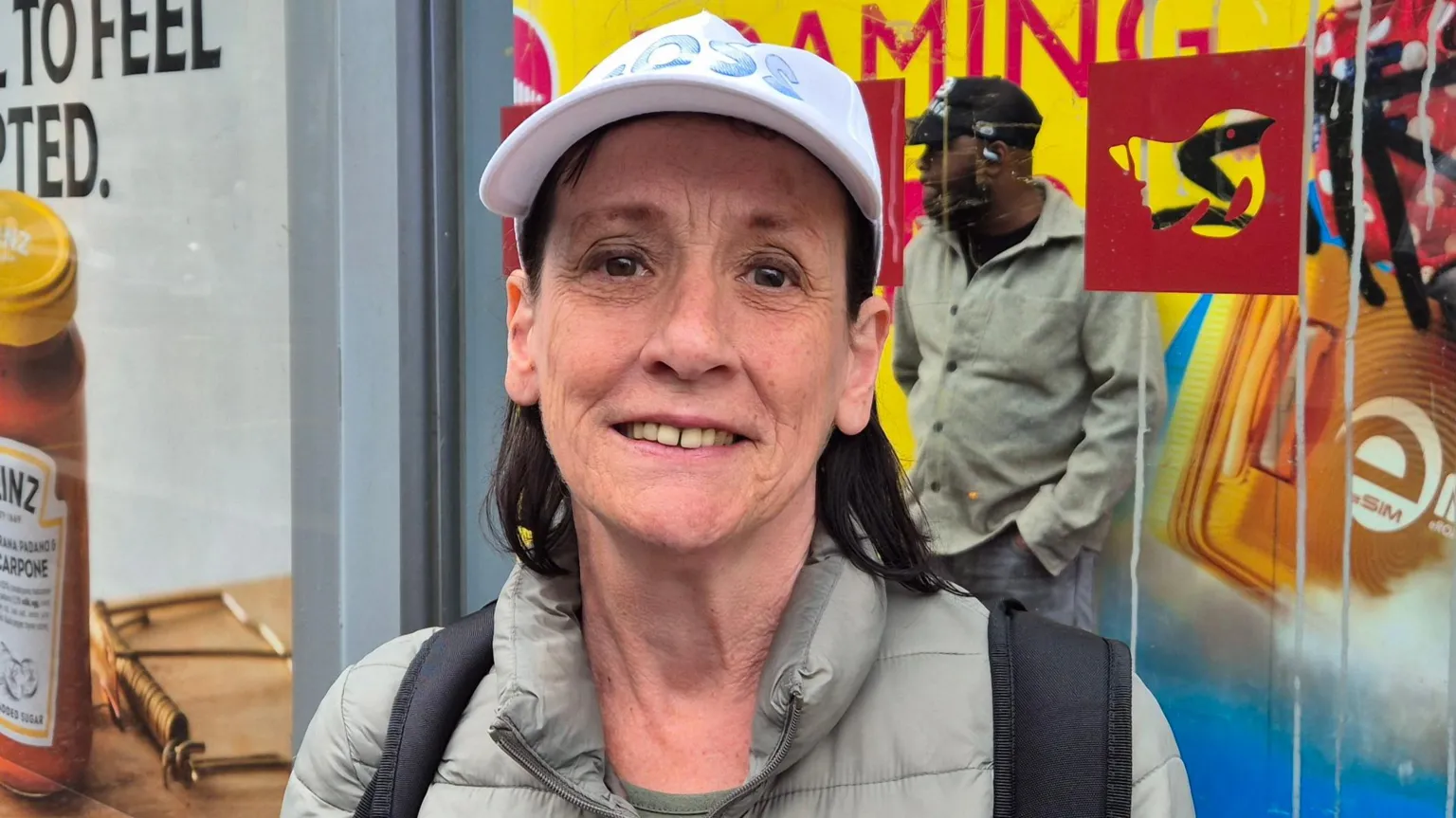 Rose Carson - a woman with short dark hair, wearing a white baseball cap. She is standing at a bus stop.