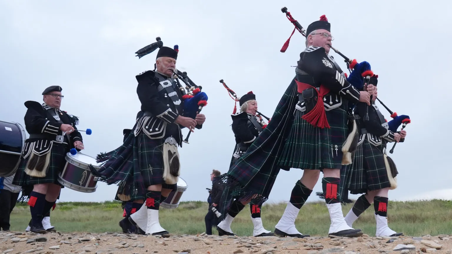  Scottish pipers walk along beach. They wear kilts and carry bagpipes and drums