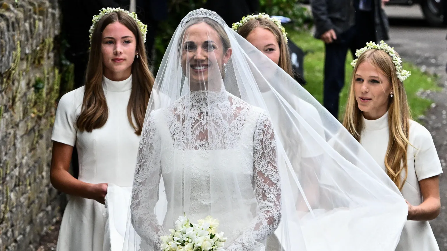  A woman is wearing a while veil and a white bride's dress, along with a tiara. She is carrying a bouquet of flowers, with three girls walking behind her holding her dress up.