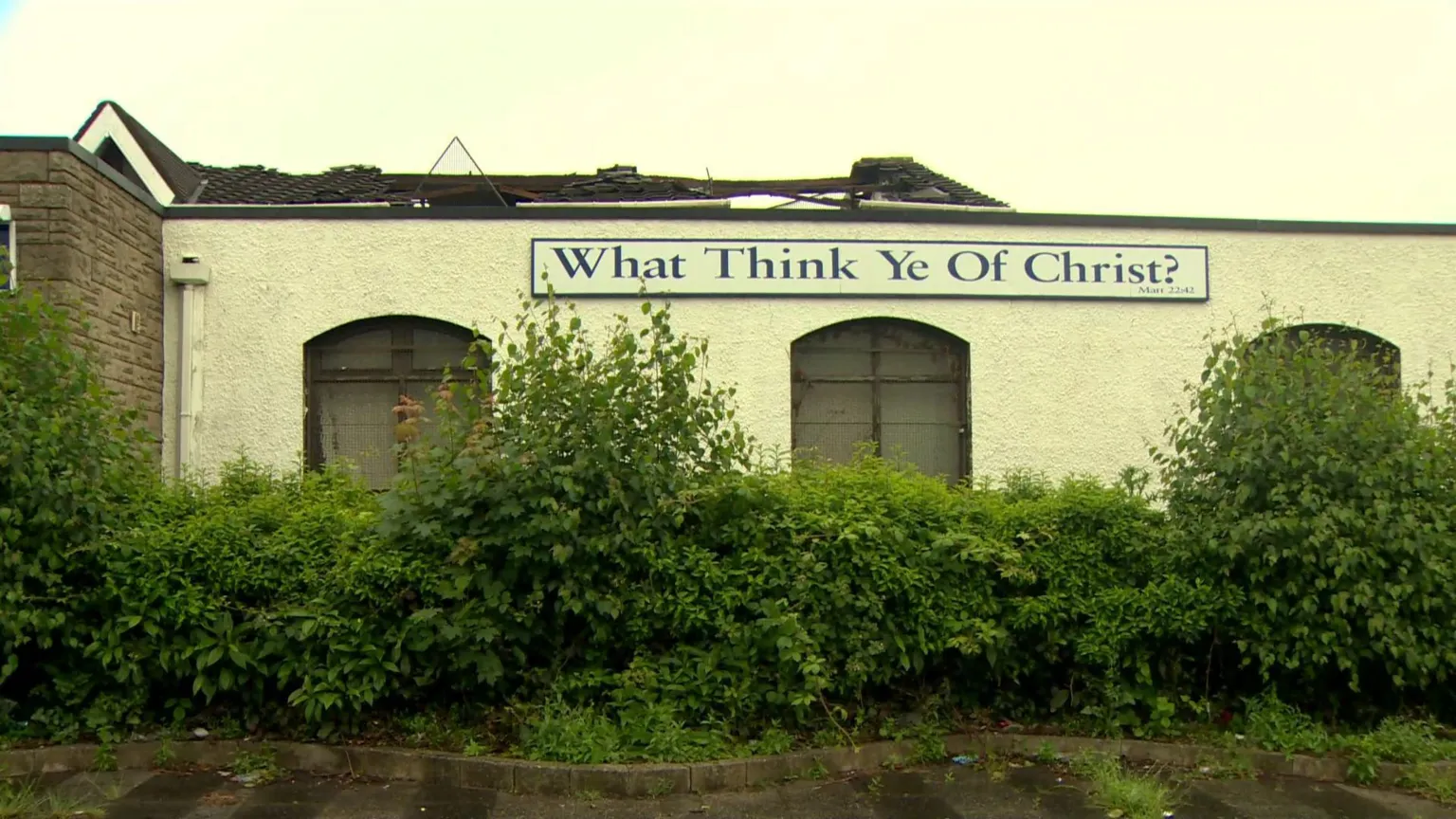 A roof of a building is damaged with sections missing. A sign on the building says, what think ye of Christ. Bushes are surrounding the building. 