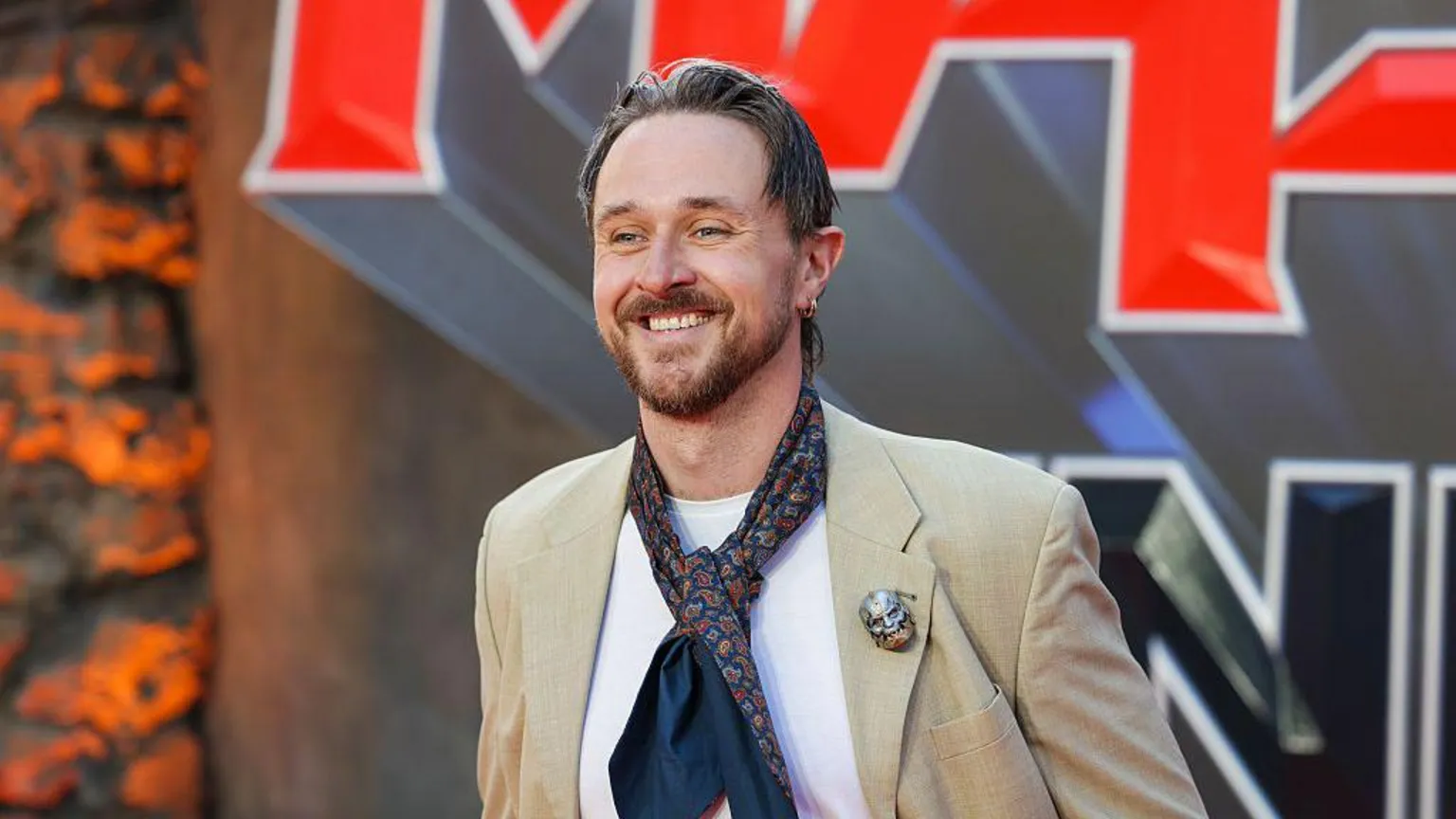  Sam Wilson at a red carpet event. He is wearing a beige suit jacket and a tie put loosely around his neck, and a white t shirt. He has light brown hair and is smiling