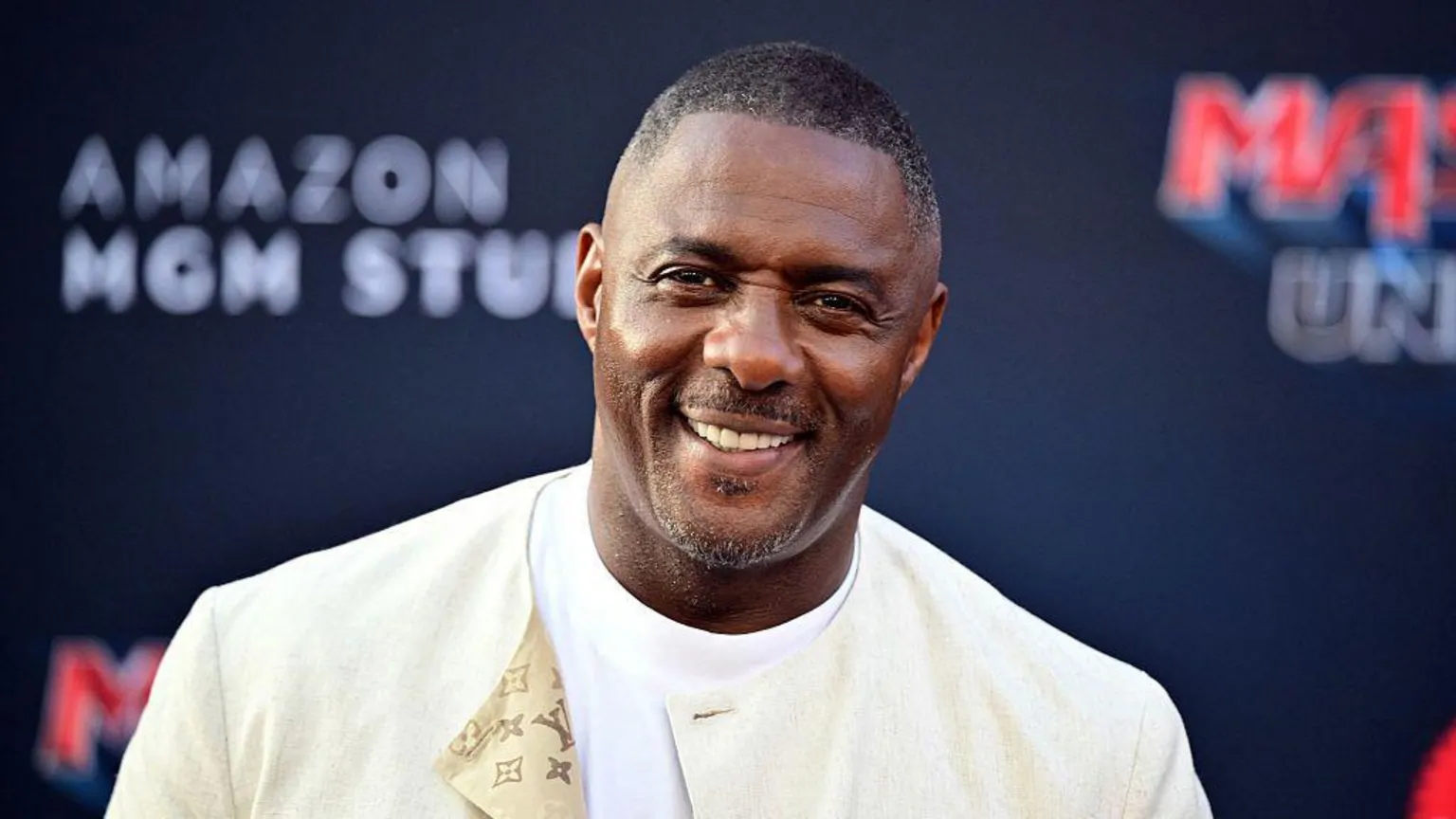  Idris Elba wearing a cream jacket over a white t-shirt. He is at a red carpet event smiling directly at the camera