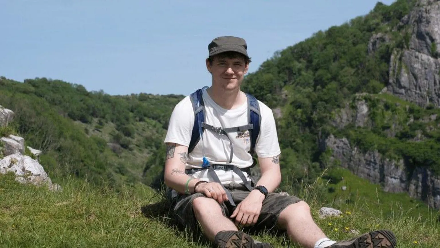 Callum Callum is wearing a white shirt and khaki shorts with a hat on. He is sat on a cliff with a rucksack on his back.