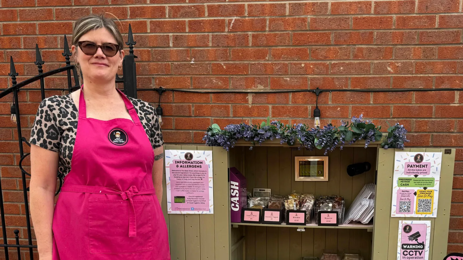 Heather Price in a pink apron and sunglasses standing next to her cake shed. The doors are open and several cakes are on display.