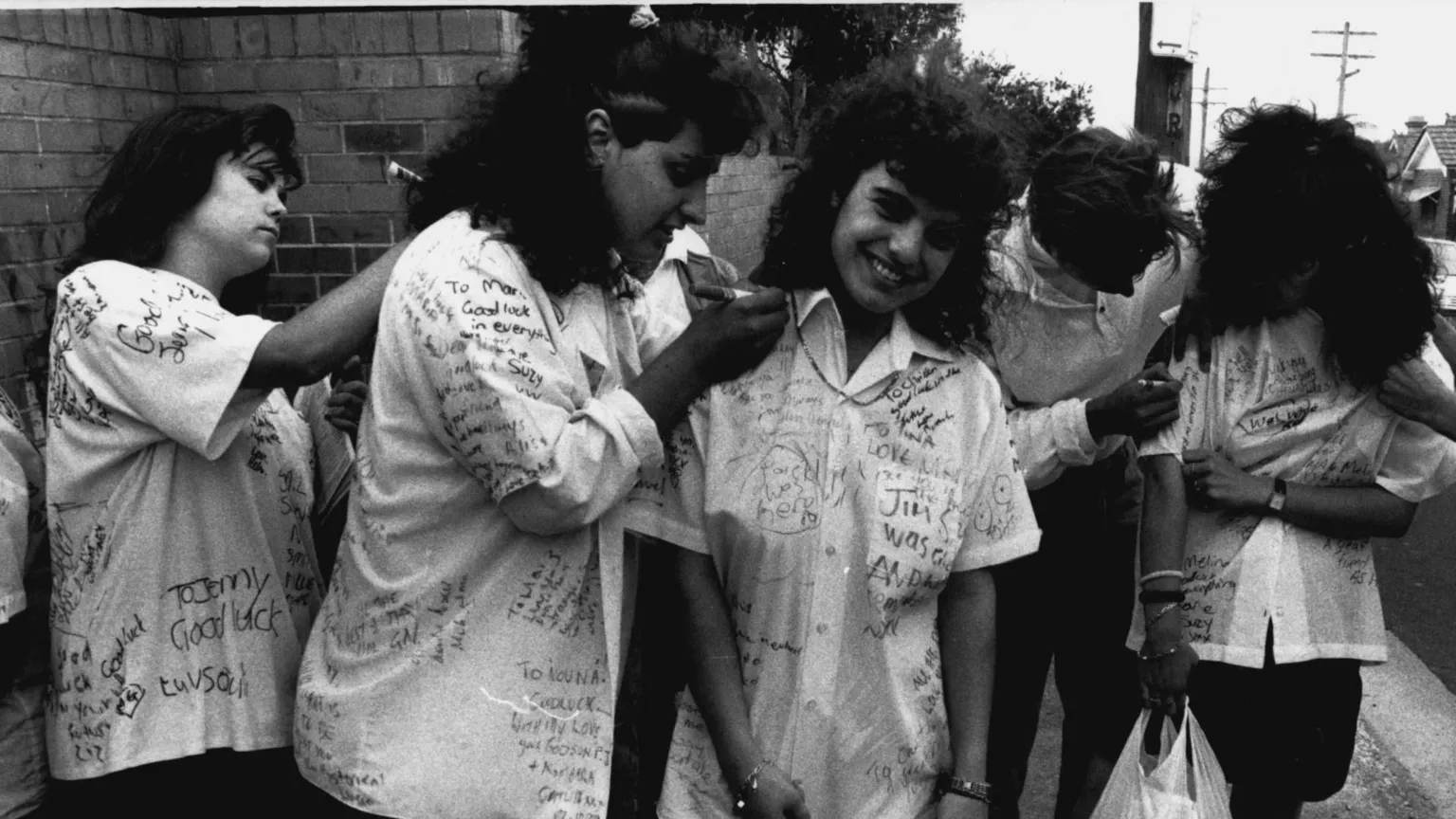  A group of students are standing outside a school getting their shirts signed with pens. 