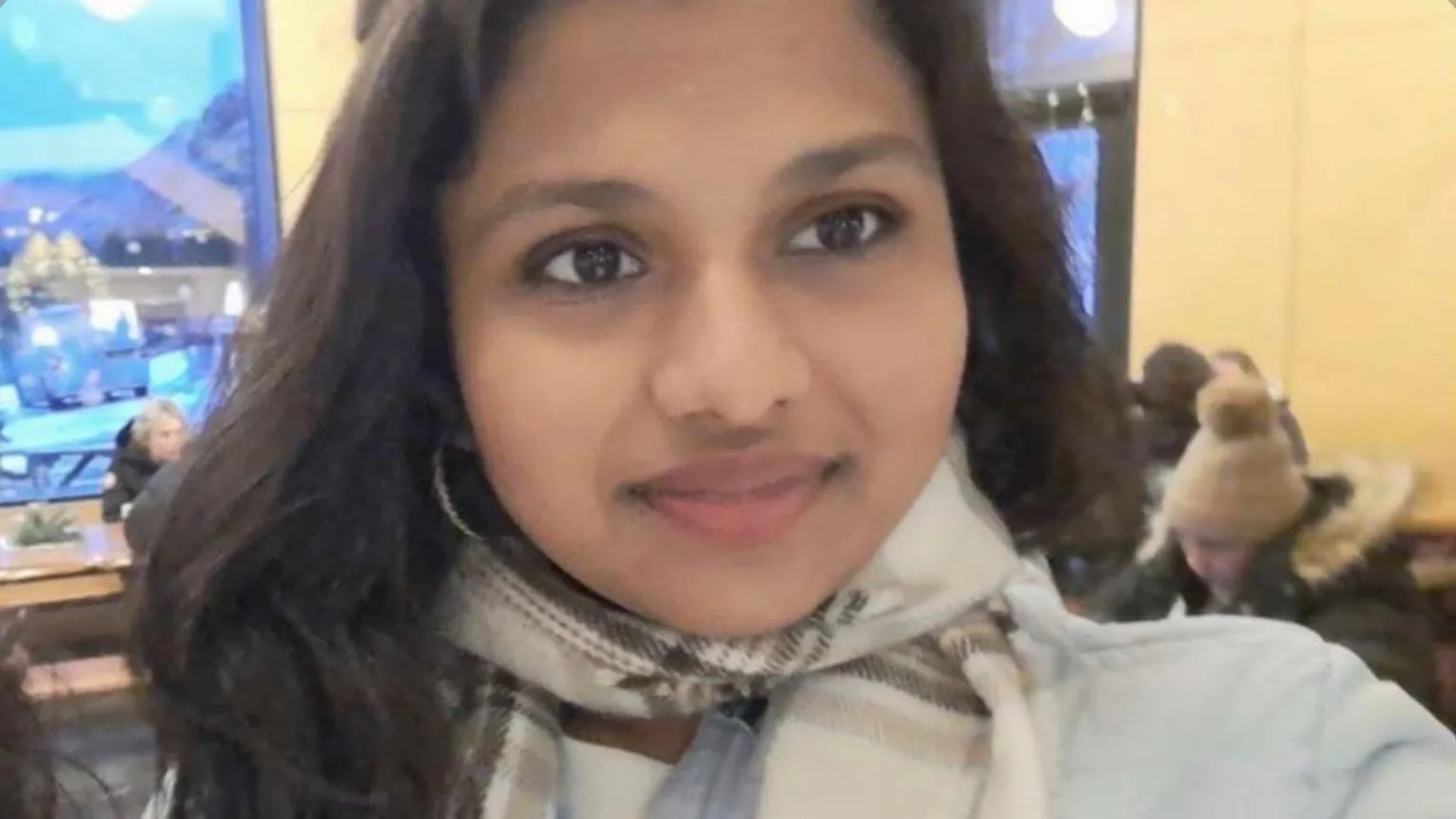 Aarushi A girl with long brown hair is wearing a checkered scarf and puffer jacket. She is standing in a restaurant smiling at the camera.