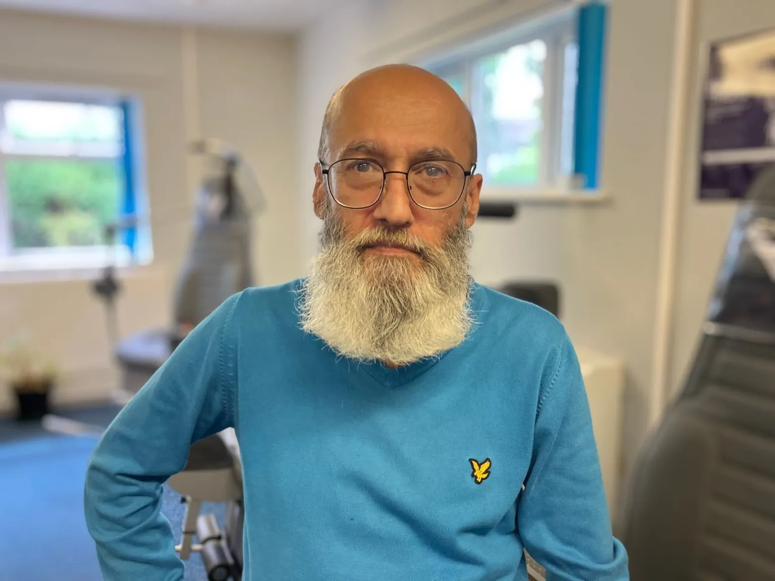 A middle-aged man with a long white beard and black glasses looks at the camera. He wears a long-sleeved blue jumper.
