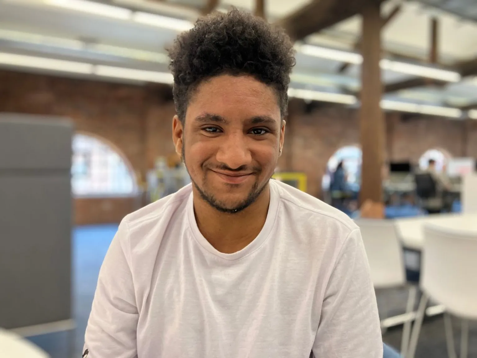 A young man in a white t-shirt smiles at the camera. The background is blurred behind him but a table and chairs are visible.
