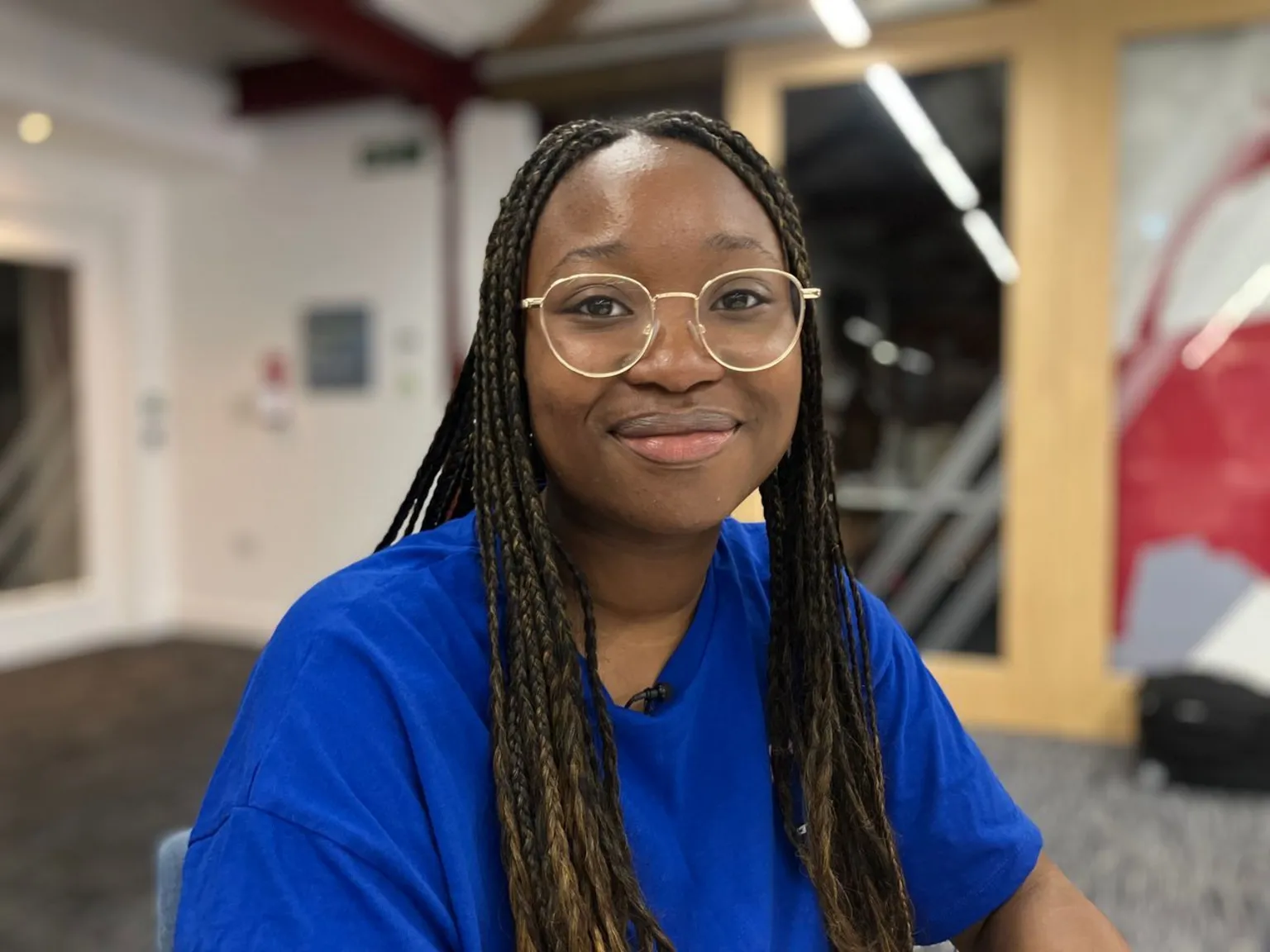 A young woman with braided hair smiles at the camera. She wears a blue t-shirt and gold glasses.
