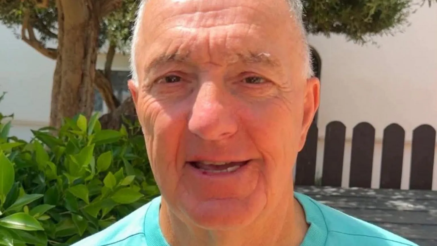 Rudi Miroli A man with grey hair and brown eyes in a light blue T-Shirt stands in front of a table and trees and a fence