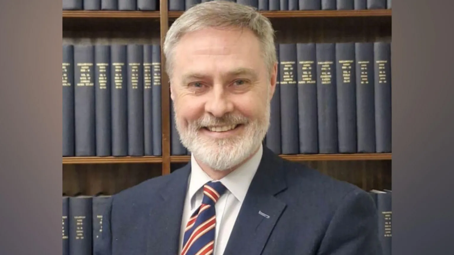 Paul Frew A man with white hair and beard, he has a navy suit, white shirt and red white and blue tie