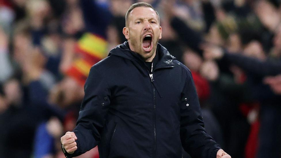Craig Bellamy in a black rain jacket roars with fists clenched
